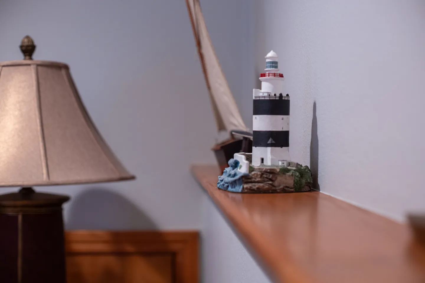 Close-up of a miniature lighthouse and model sailboat displayed on a wooden shelf next to a table lamp.