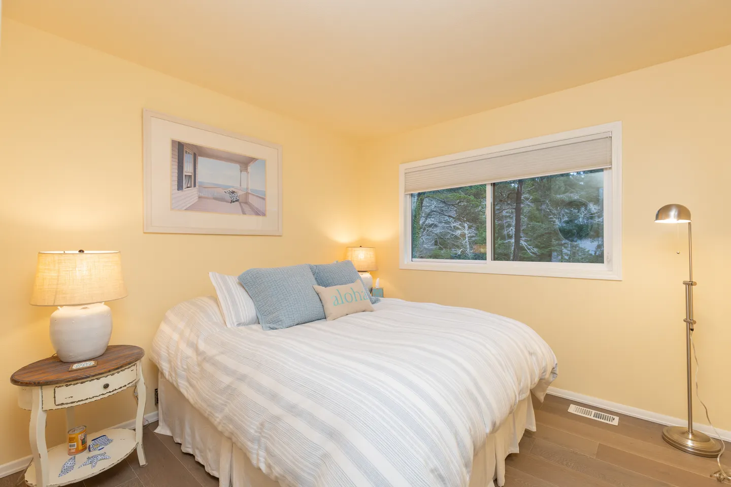 Bedroom with yellow walls, a made bed with striped bedding, a small bedside table, a floor lamp, and a window showing outdoor greenery.