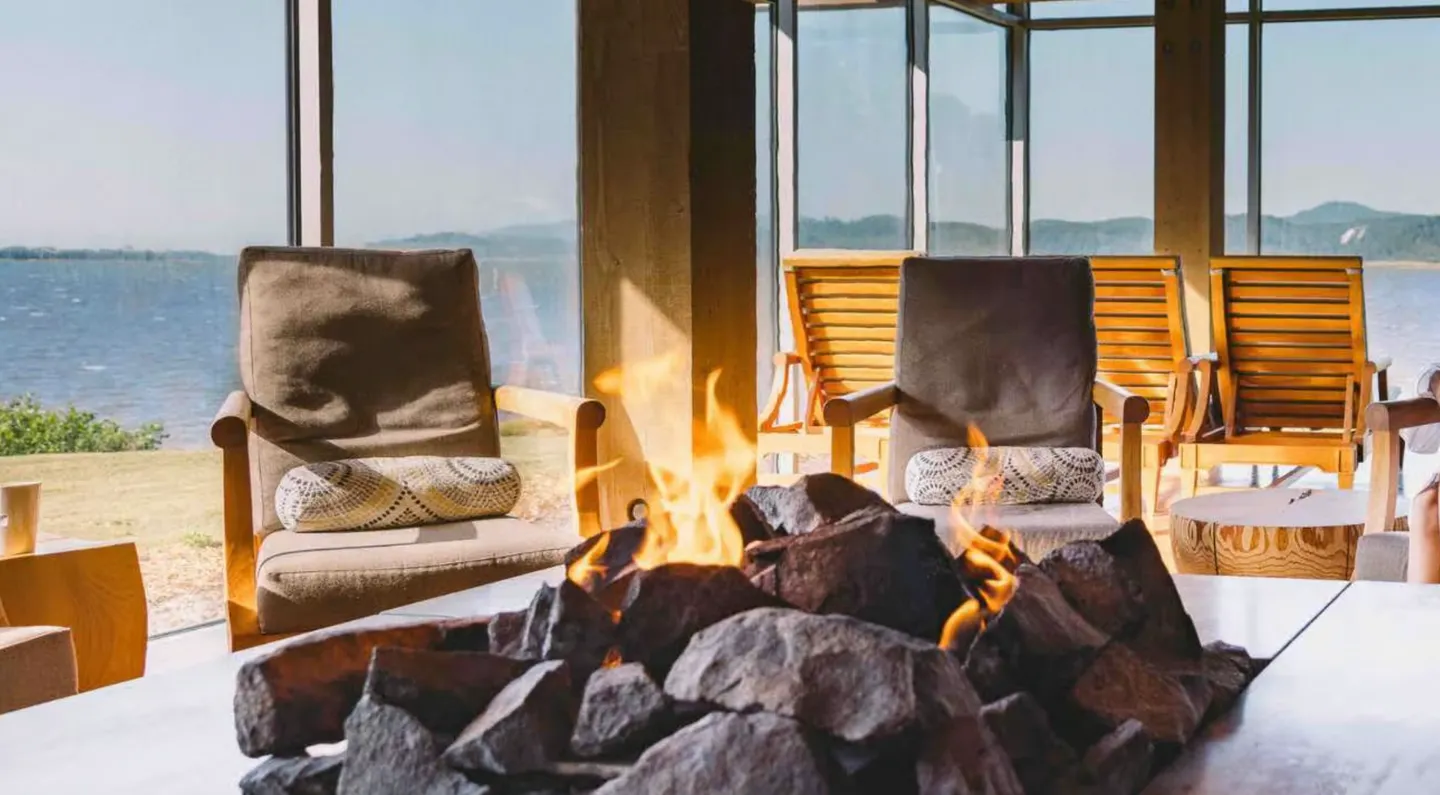 Indoor seating area with an active stone fire pit feature looking through glass walls out toward the water.
