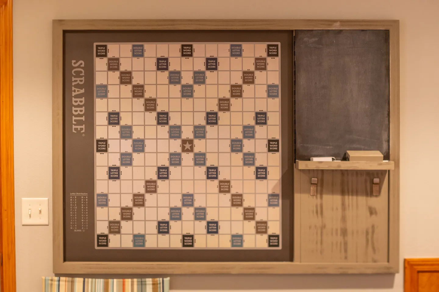 A wall-mounted Scrabble board framed in wood with an attached chalkboard and scorekeeper shelf.
