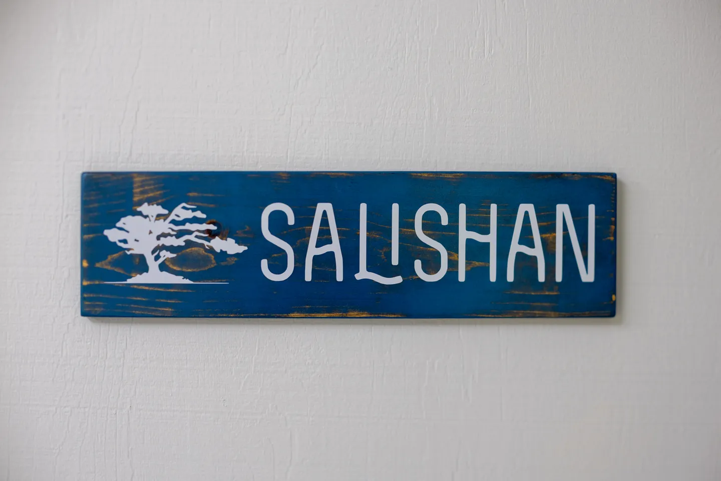 A rectangular blue wooden sign mounted on a textured off-white wall reading 'SALISHAN' with a white tree emblem on the left.