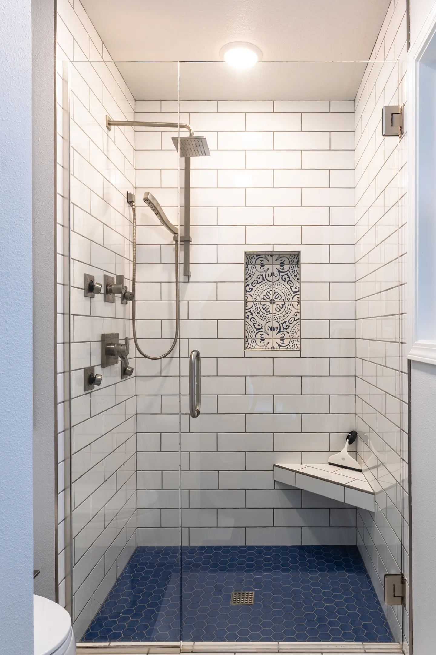 Walk-in shower with glass enclosure, white subway tile walls, blue hexagonal floor tile, dual showerheads, and a built-in corner bench.