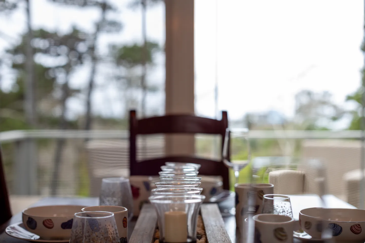 Close-up of a set dining table with bowls, glasses, and candle holders in front of large windows.