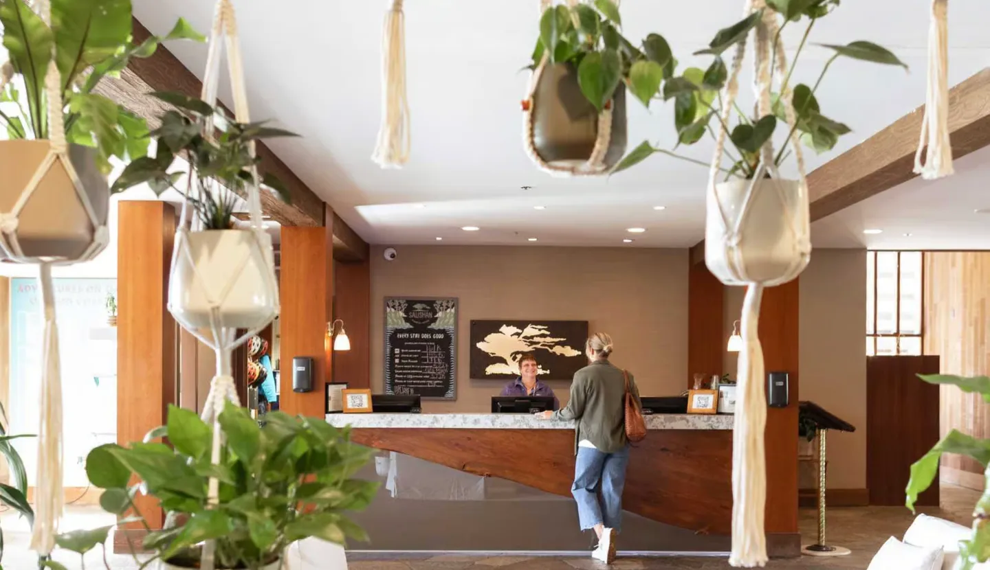 Reception desk in a lobby viewed past hanging macramé planters, with a guest speaking to front desk staff.