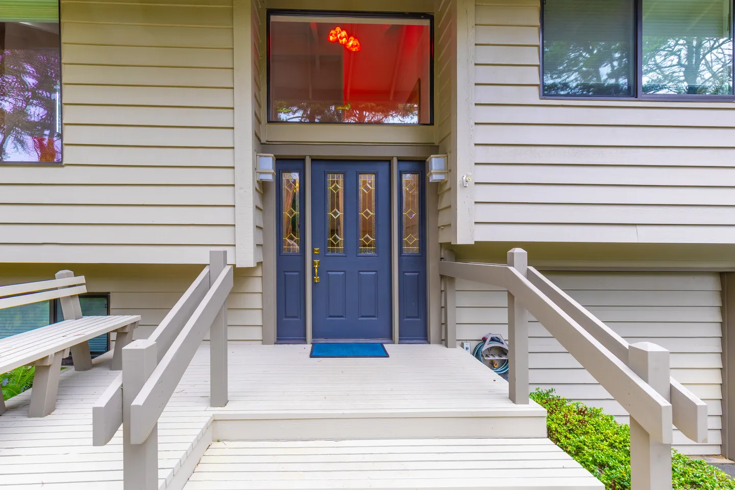 Front entrance of a house featuring light-colored siding, a wooden deck with railings, a built-in bench, and a blue front door with glass sidelights.