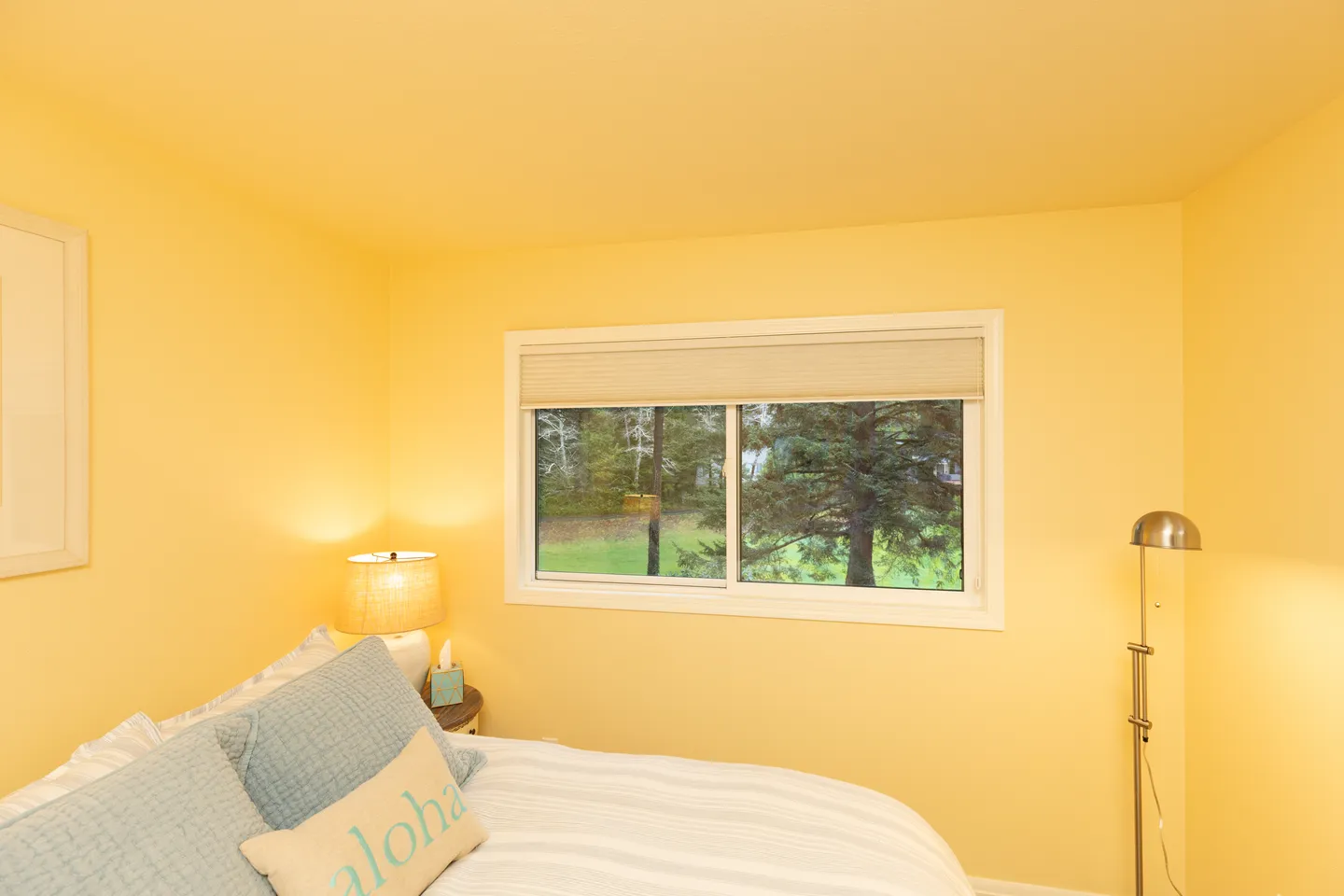 Bedroom with bright yellow walls, a window overlooking trees, a bed with blue pillows, and bedside lighting.