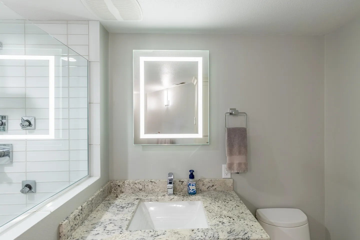 Bathroom vanity with a speckled countertop, undermount sink, backlit LED mirror, and a glass-enclosed shower on the left.