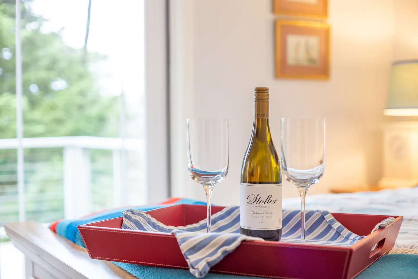 Close-up of a red leather tray holding two wine glasses and a bottle of white wine on a bed by a bright window.