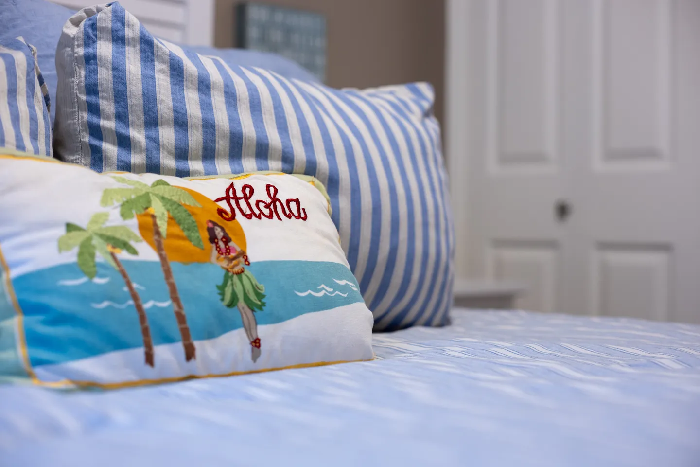 Close-up of a bed with blue-and-white striped pillows and an embroidered tropical throw pillow reading Aloha.