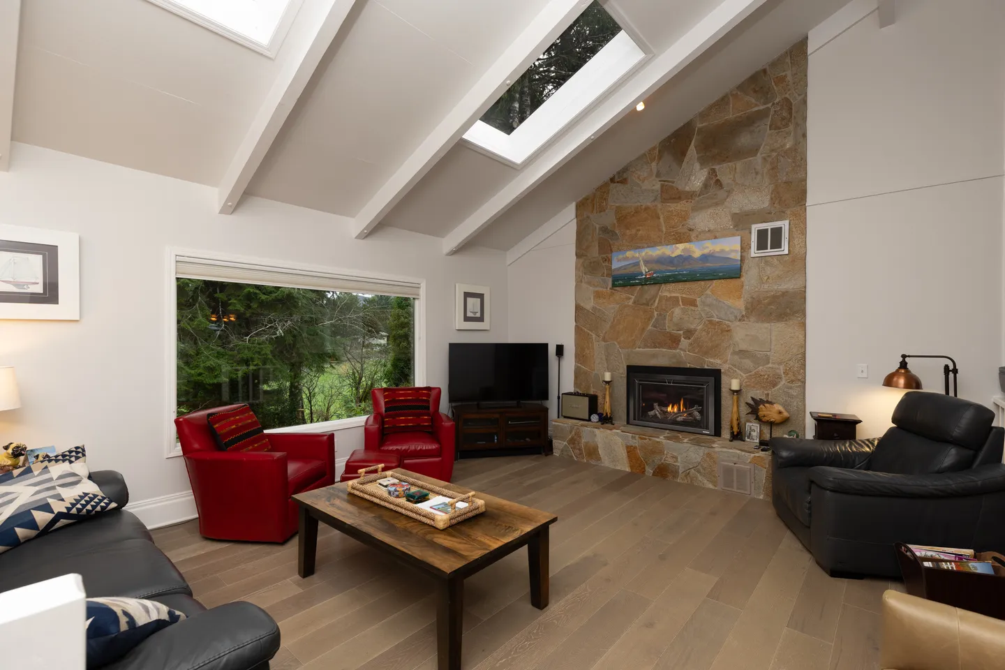 Living room with vaulted beamed ceilings, skylights, stone fireplace, red and black armchairs, and a large window.