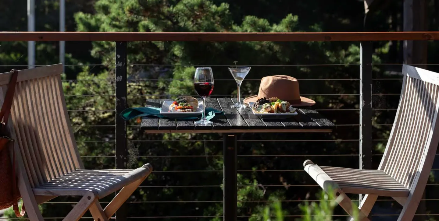 A small outdoor table with two chairs, drinks, and food set against a deck railing overlooking green trees.