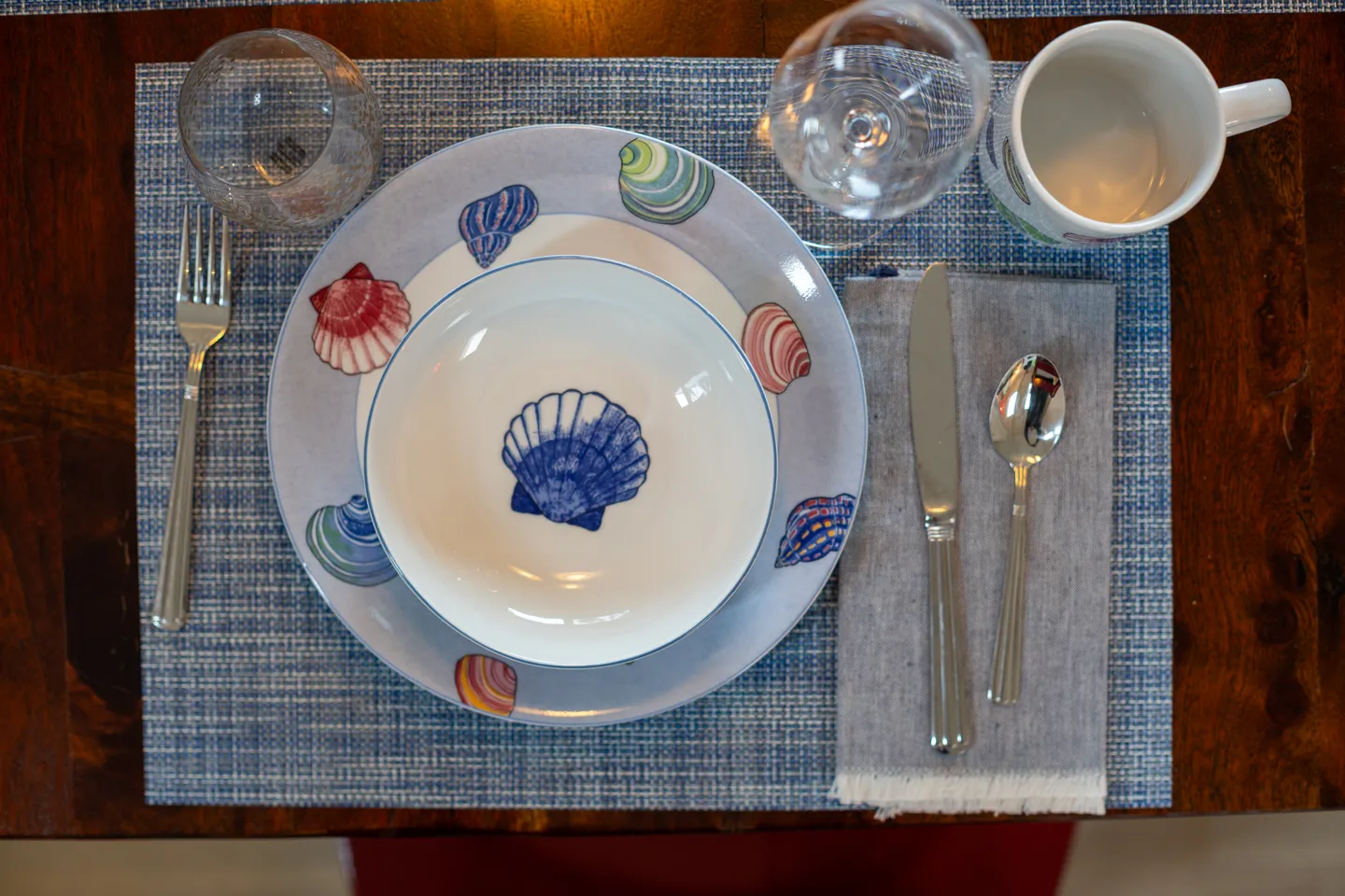 Overhead view of a dining place setting with seashell-patterned dinnerware, flatware, glasses, and a mug on a woven placemat.
