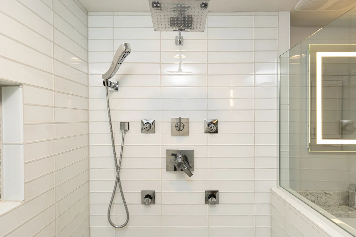 Close-up of a tiled shower stall featuring horizontal white subway tiles, a rain showerhead, a handheld sprayer, body jets, and modern chrome controls.
