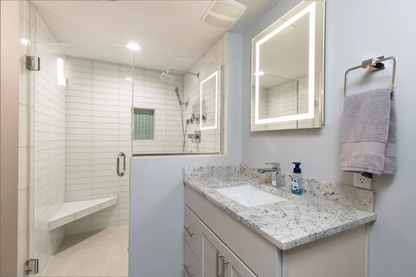Bathroom with a vanity featuring a speckled countertop and an illuminated mirror next to a walk-in shower with white tile and a bench.