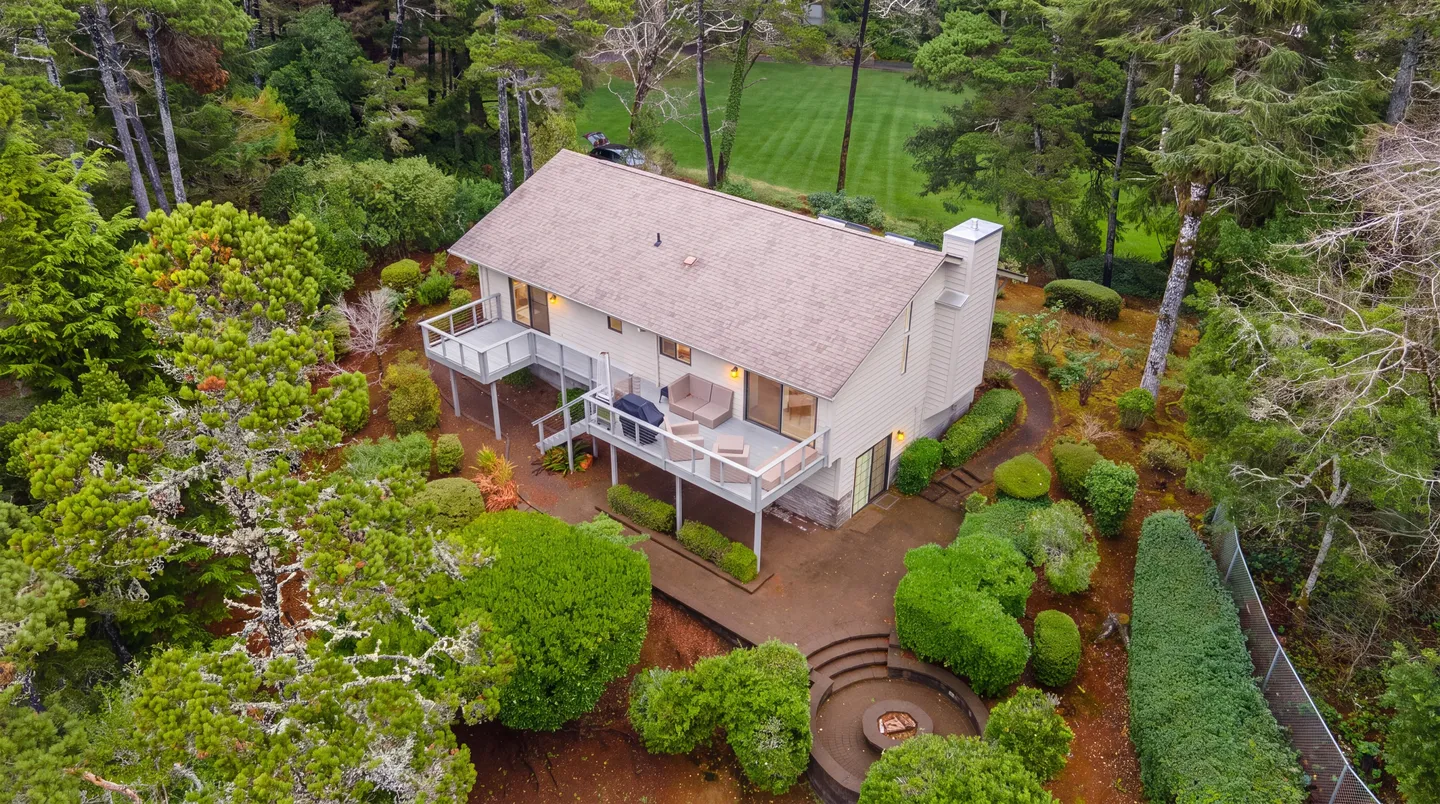 Aerial view of a two-story home surrounded by tall trees and manicured shrubs, featuring an elevated deck and a sunken fire pit.