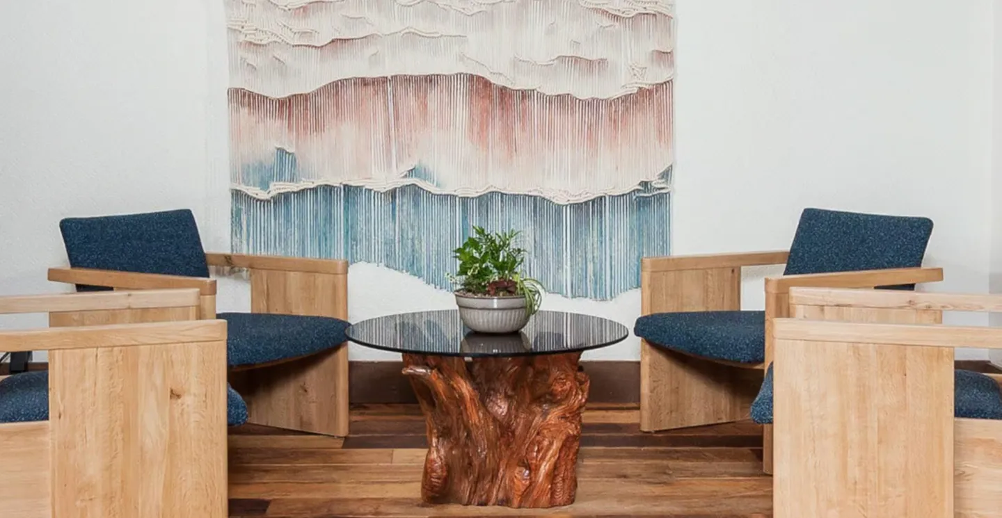 Seating area featuring wooden armchairs with blue upholstery surrounding a glass-topped tree root table under a wall tapestry.