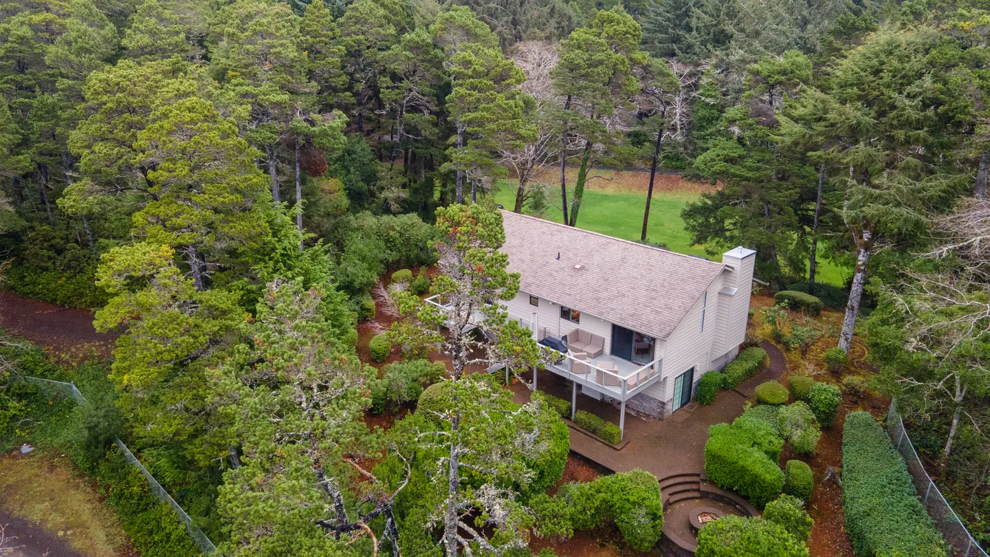 Aerial view of a two-story home surrounded by dense evergreen trees with a deck and landscaped yard.