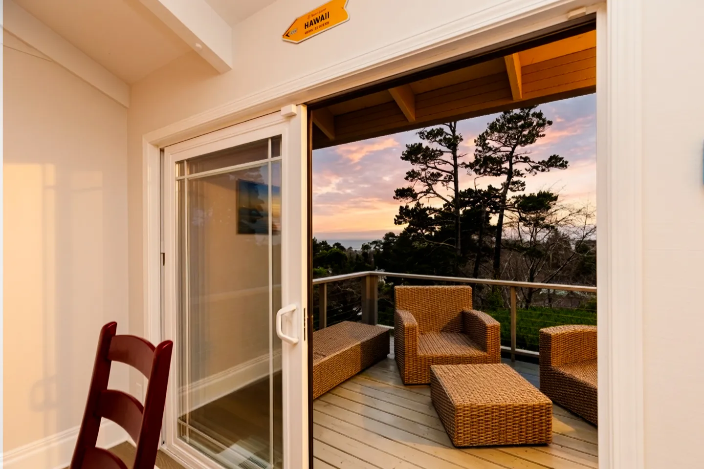Open sliding glass door looking out from an indoor room onto a wooden balcony with woven patio seating at sunset.