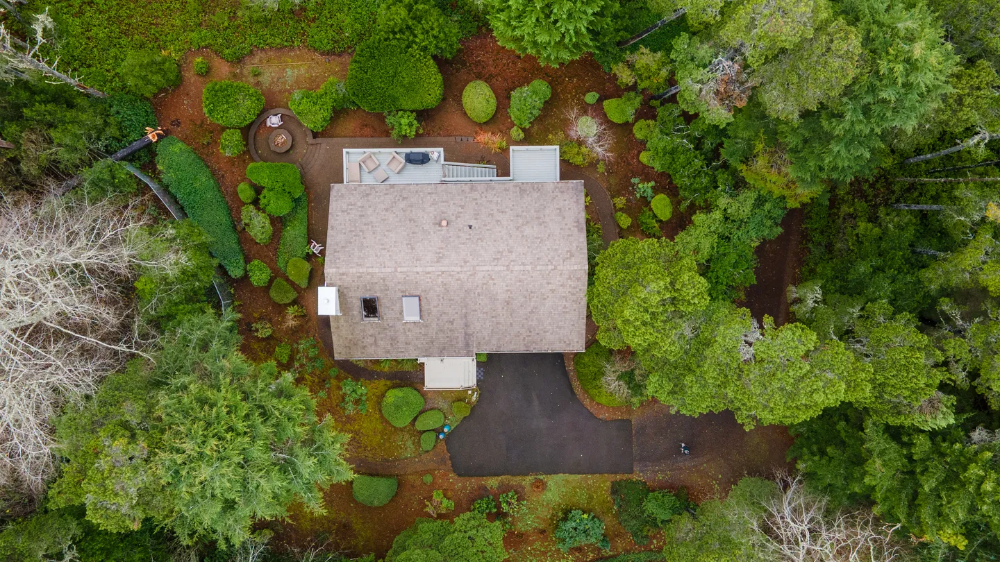 Overhead aerial view of a house nestled in a dense forest with a paved driveway, patio, and landscaped shrubs.