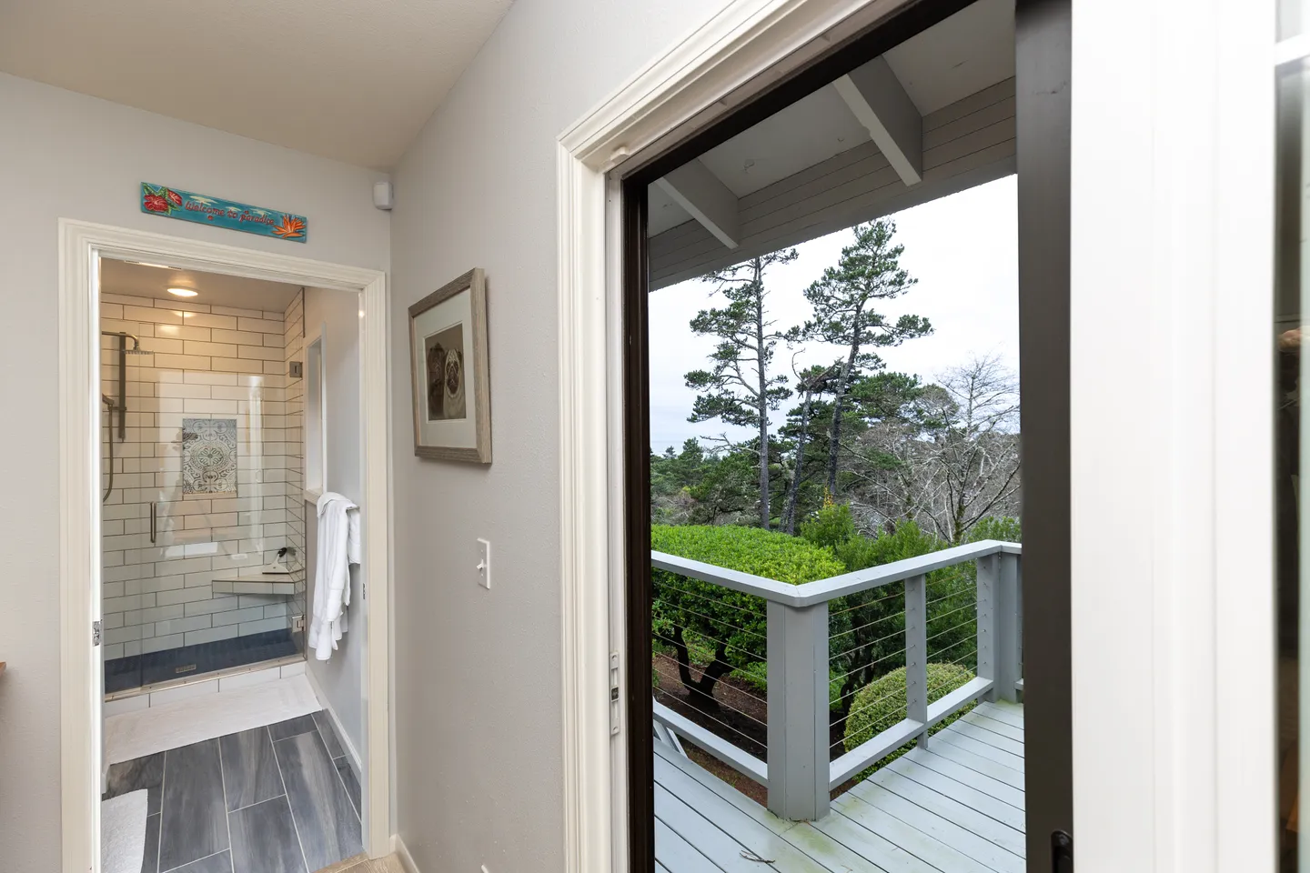 Open sliding glass door leading to a balcony surrounded by trees, adjacent to a hallway with an open doorway to a tiled bathroom.