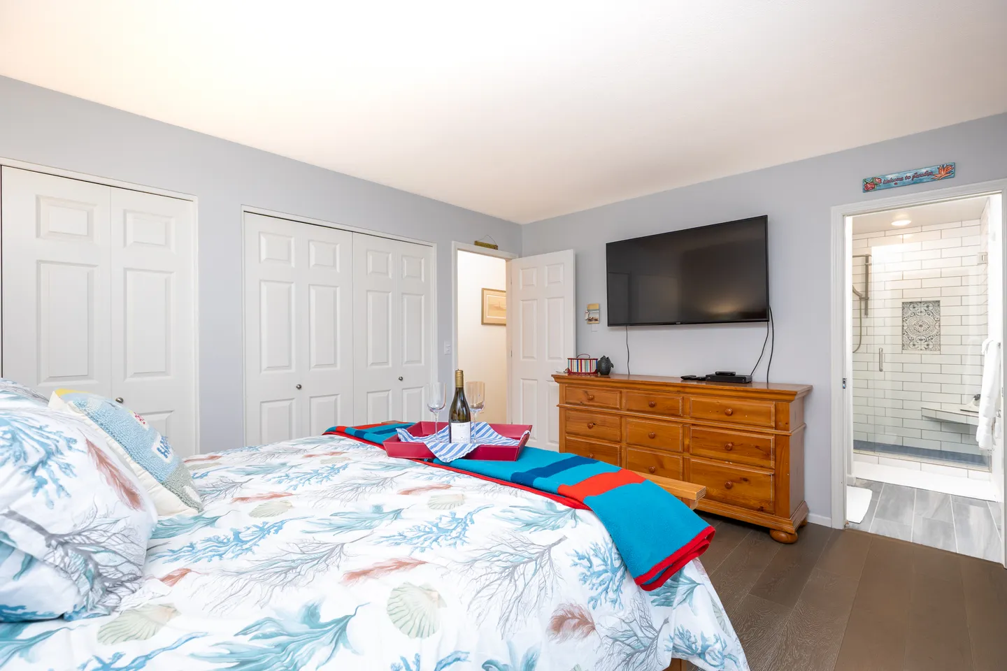 Bedroom featuring a coastal-themed bedspread, a wooden dresser beneath a wall-mounted TV, closet doors, and an open doorway into an ensuite bathroom.