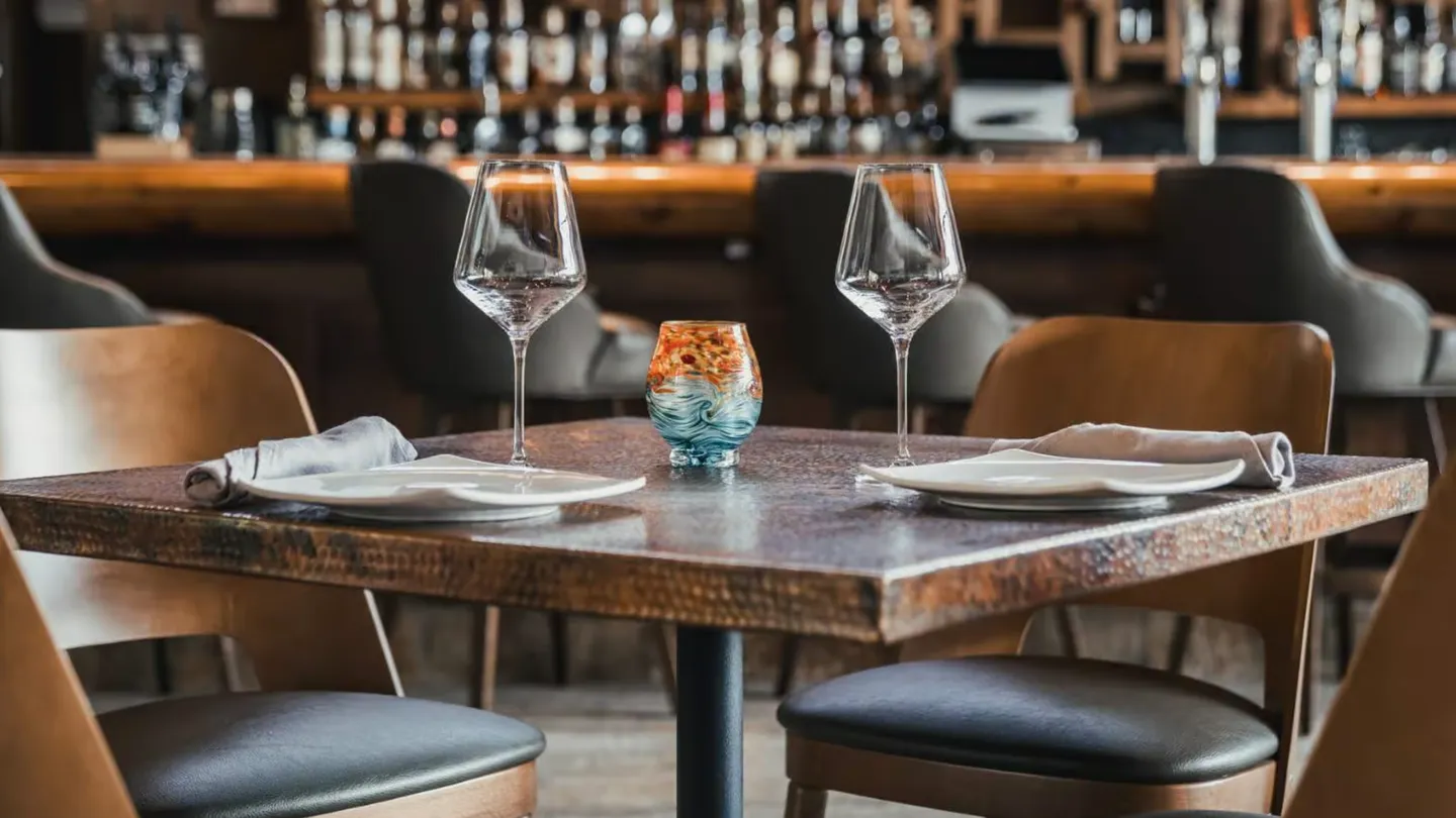 Close-up of a square dining table set with two wine glasses, plates, and a decorative candleholder, with bar seating in the blurred background.