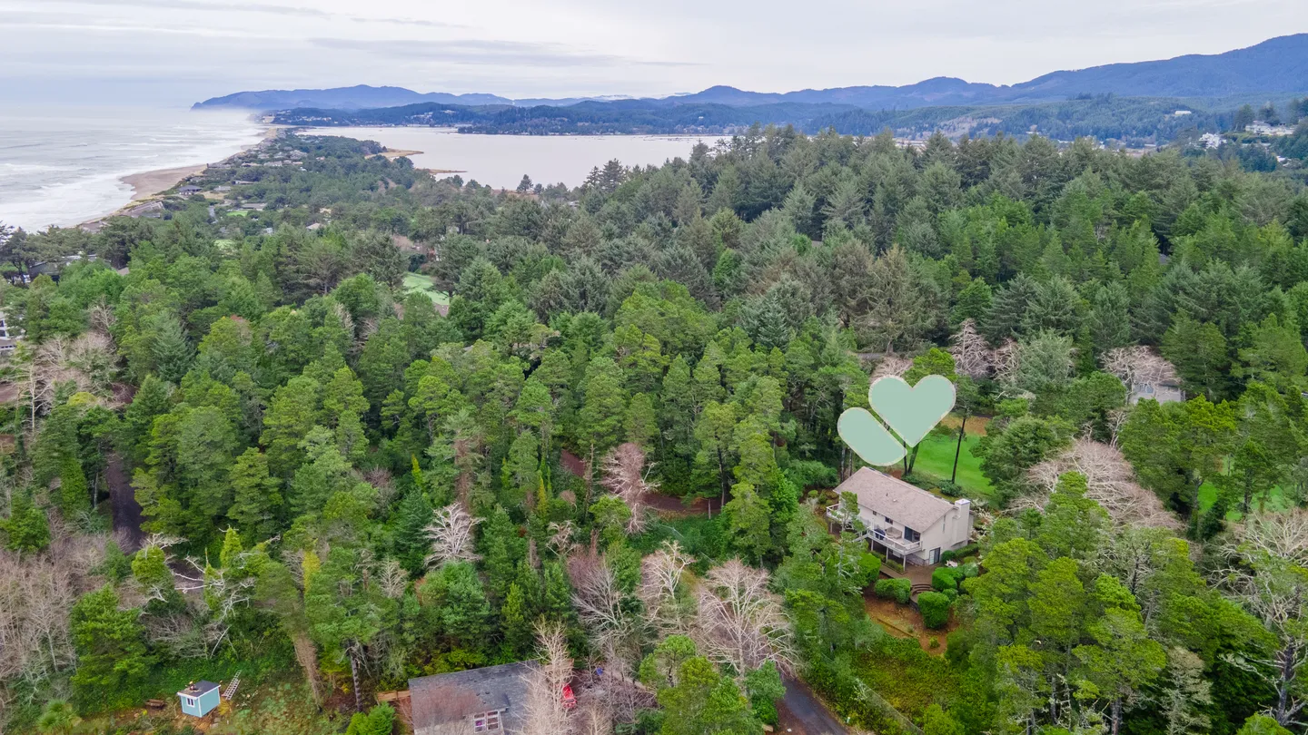 Aerial view of a coastal house nestled in dense evergreen forest overlooking a bay and ocean coastline.