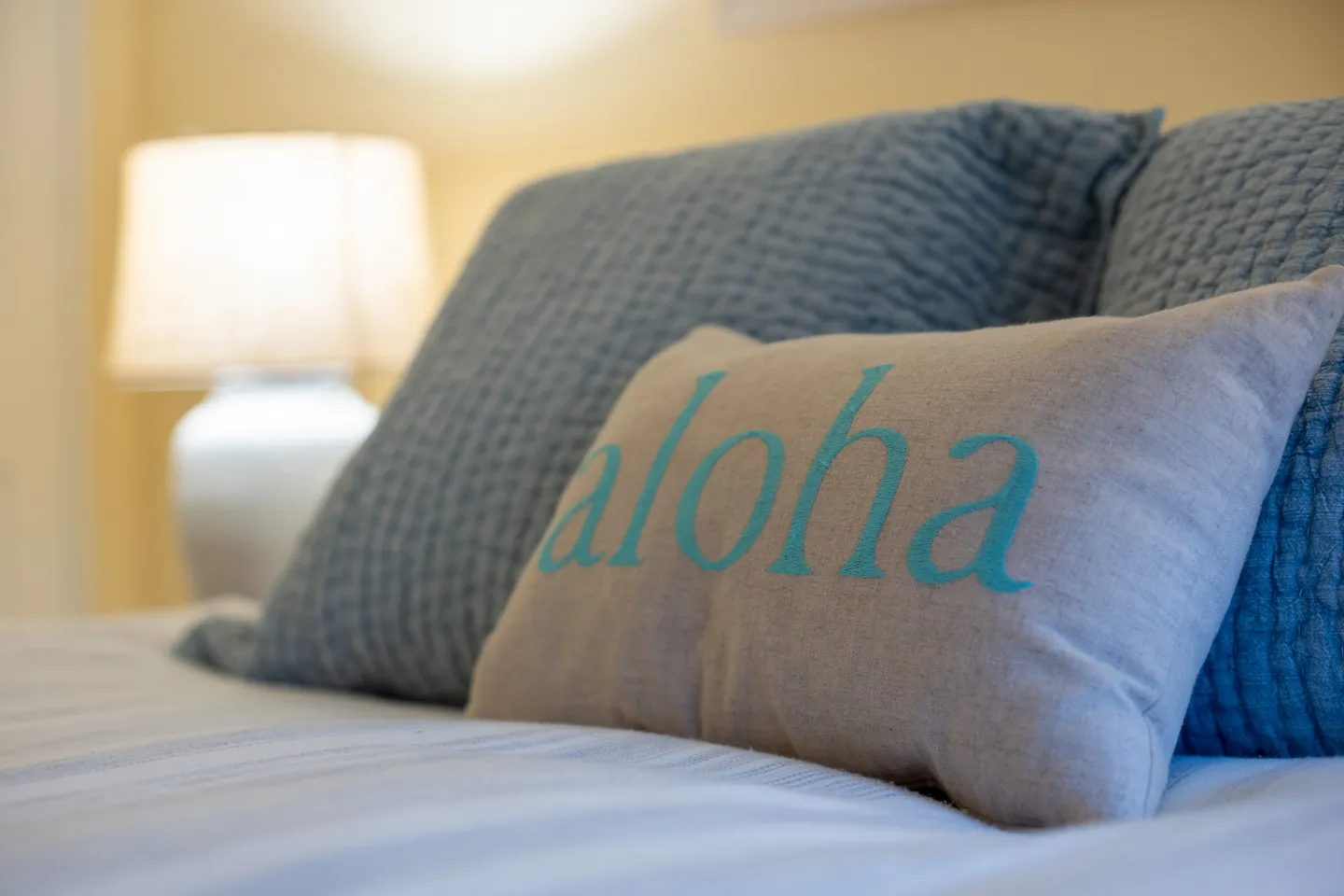 Close-up of a bed with a decorative pillow that reads 'aloha' in blue lettering, alongside blue textured pillows and an illuminated bedside lamp.