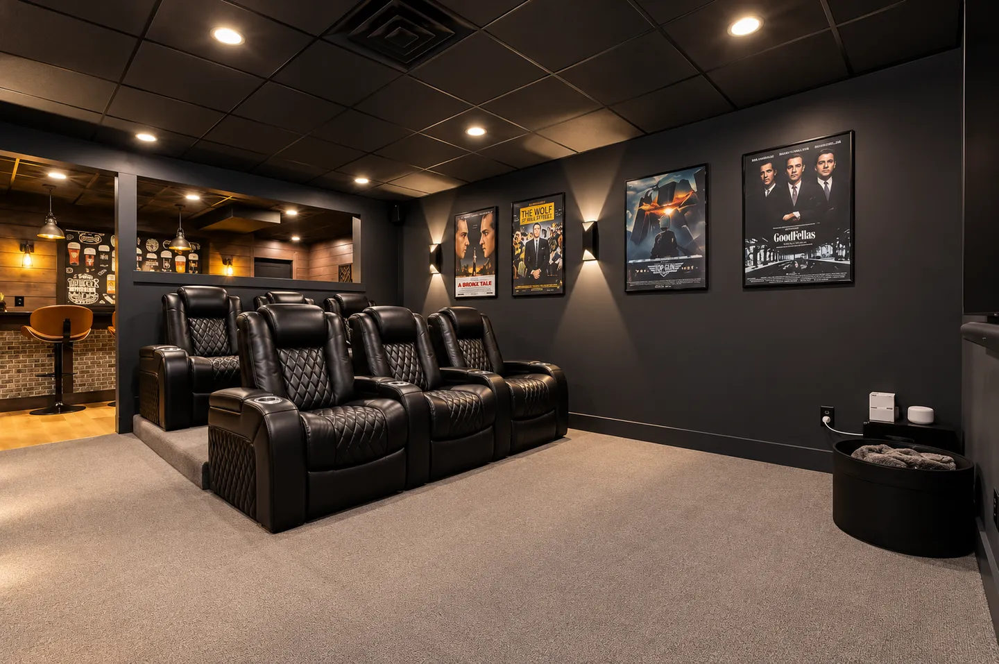 A home theater room featuring two tiered rows of black leather recliner seats, framed movie posters on dark walls, and a cutout looking into an adjacent bar area.