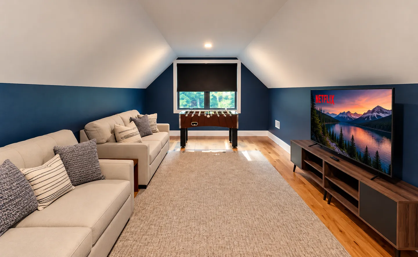 Upstairs lounge and game room with sloped ceilings, two light-colored couches, a television on a console, and a foosball table under a window.