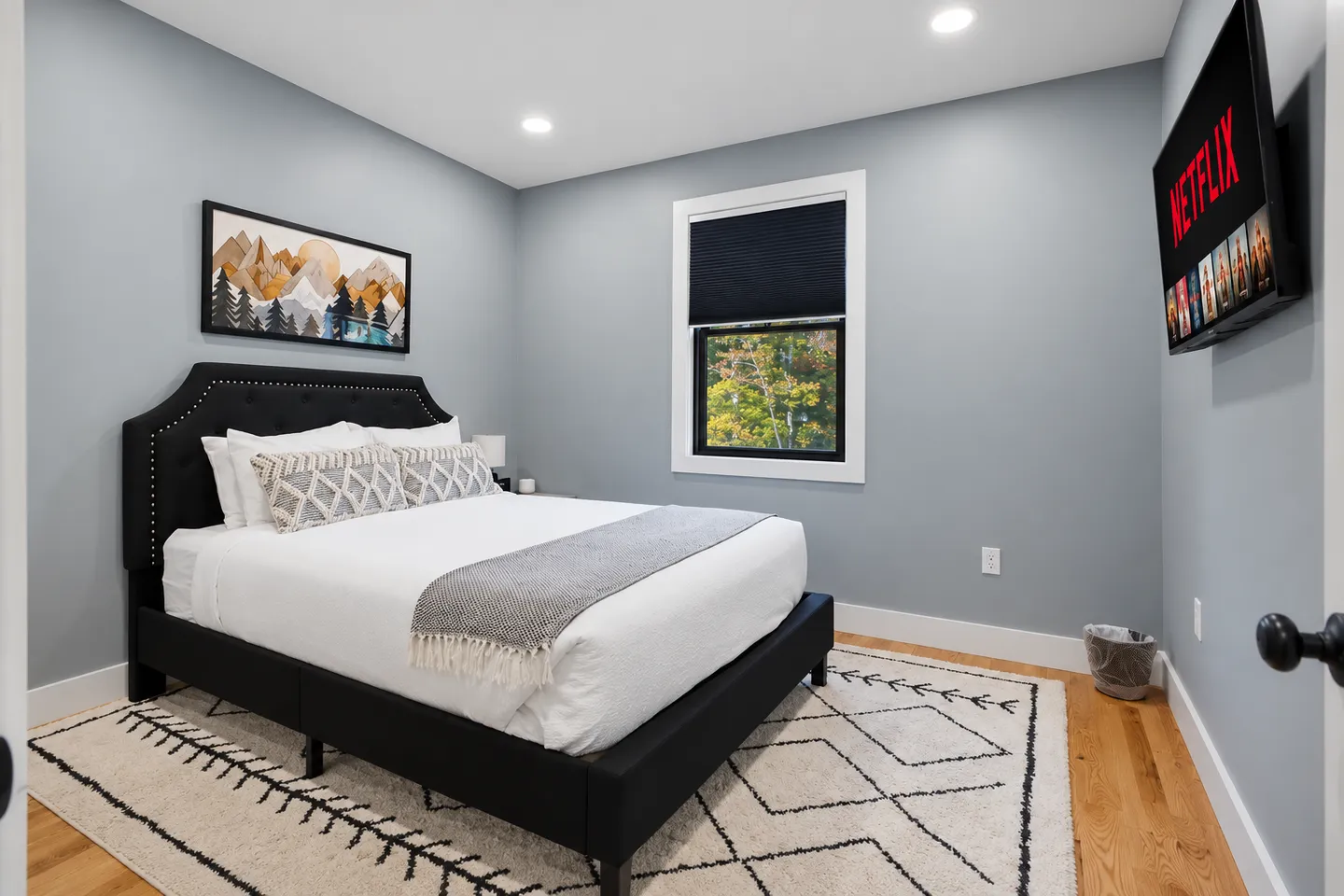 Bedroom with a black upholstered bed, light blue walls, geometric area rug, window, and wall-mounted television.