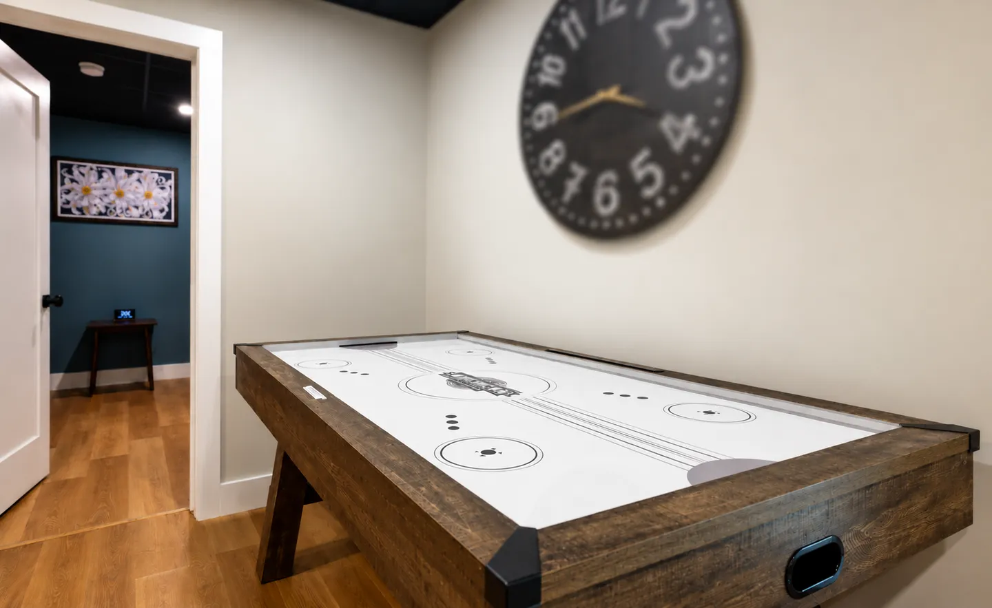 An air hockey table sits against a neutral wall beneath a large round wall clock, with an open doorway leading to an adjoining room.