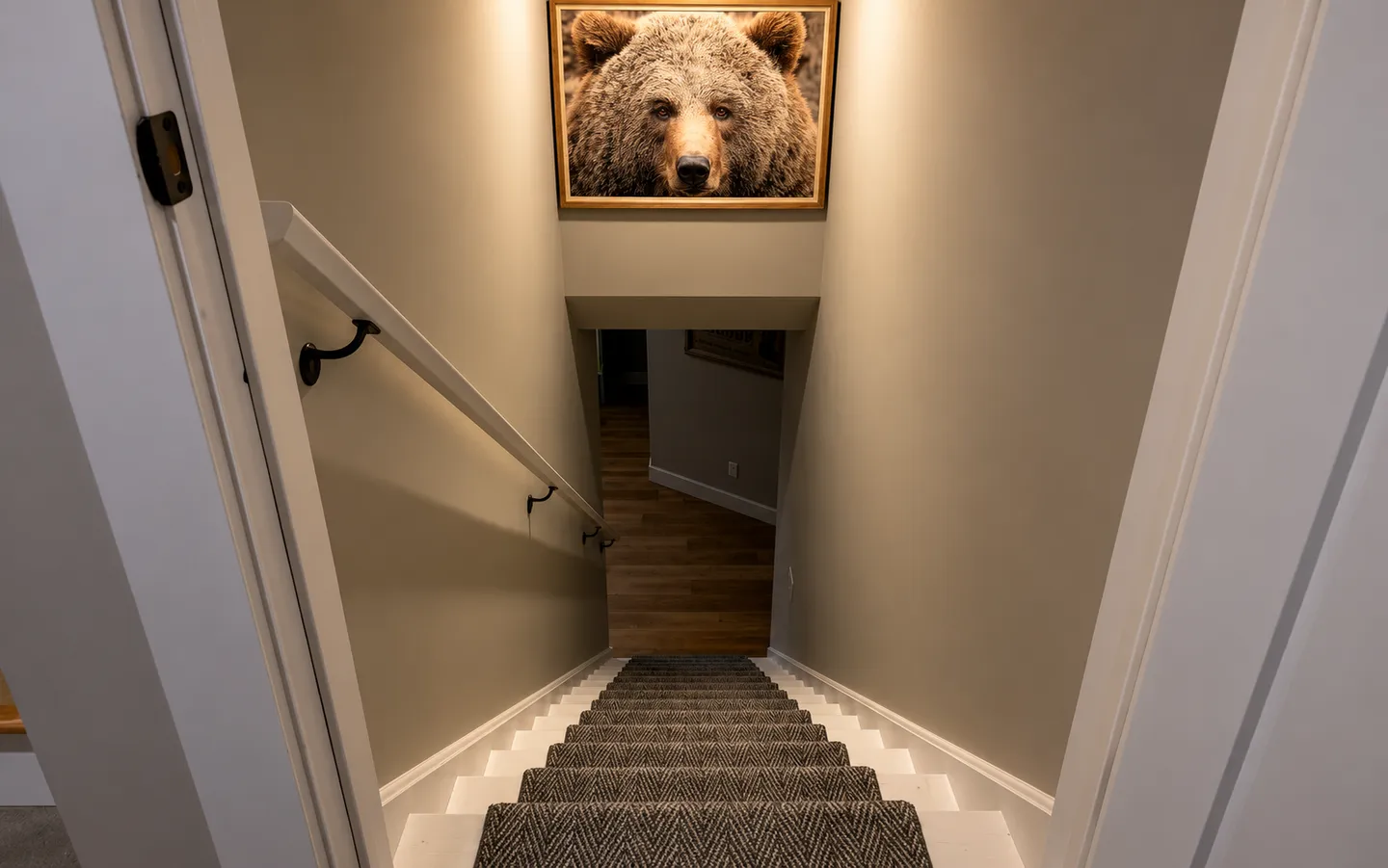 Looking down a carpeted staircase with a white handrail toward a lower level, beneath a framed picture of a brown bear.