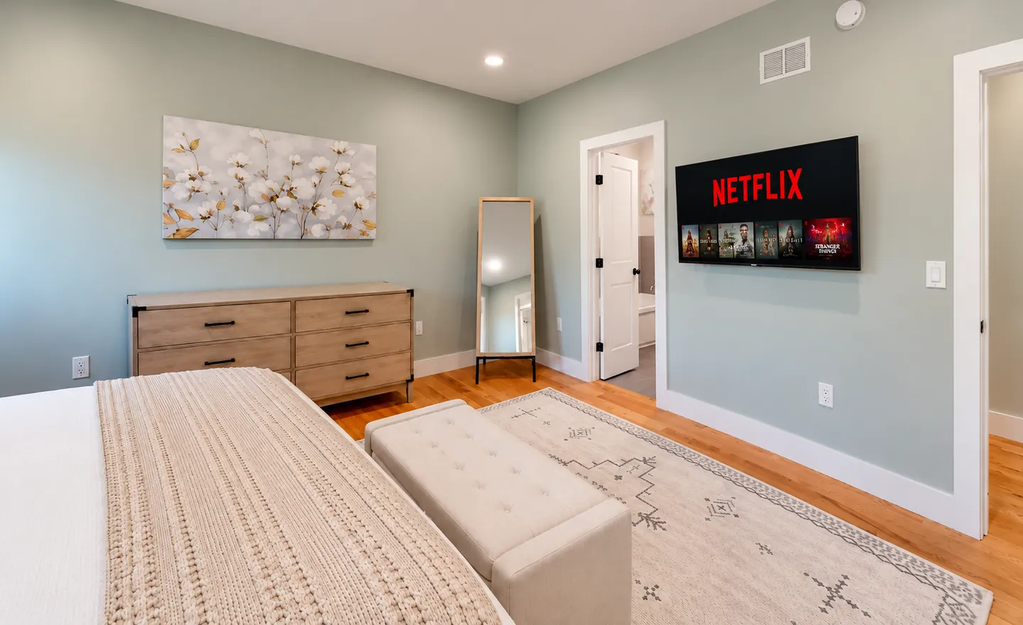 Bedroom with light green walls, a wall-mounted flat-screen TV, a wooden dresser, a full-length mirror, and wood flooring.