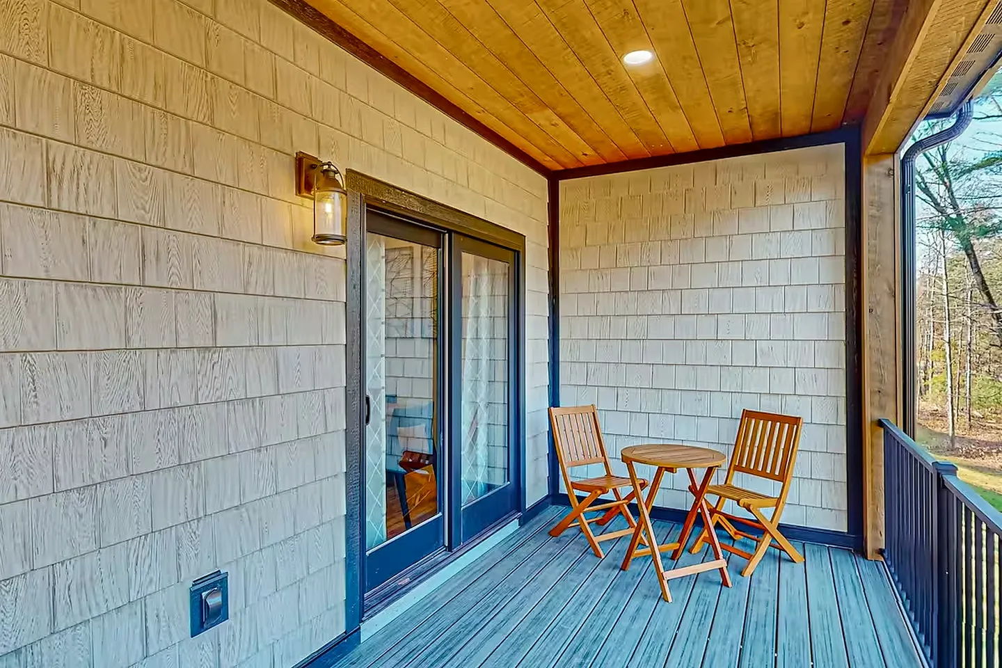 Covered deck with light shake siding, wood plank ceiling, sliding glass doors, and a small wooden bistro table with two folding chairs.