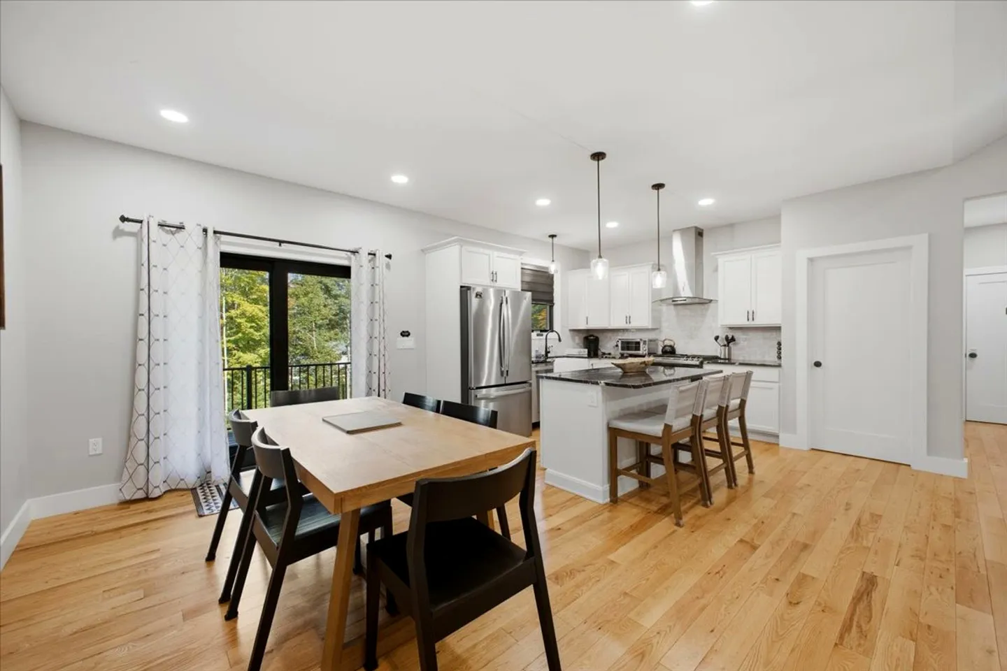 Open-concept dining area and kitchen with light wood flooring, a dining table with chairs, an island with barstools, white cabinets, and sliding glass doors to a balcony.
