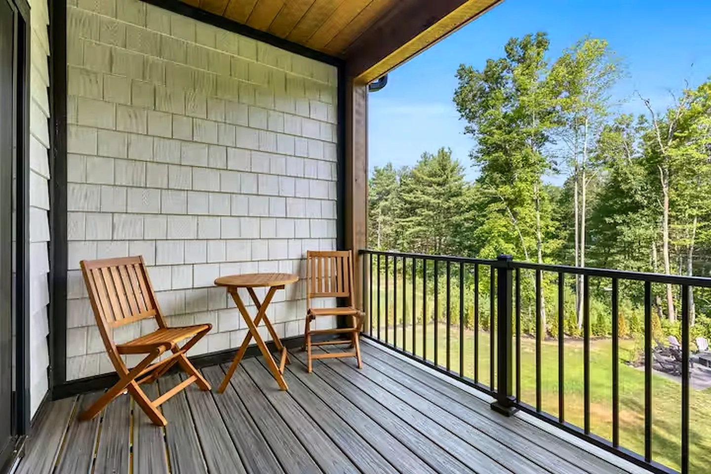 A covered deck with wood-style flooring, a railing, and two wooden folding chairs around a small round table, overlooking a wooded yard.