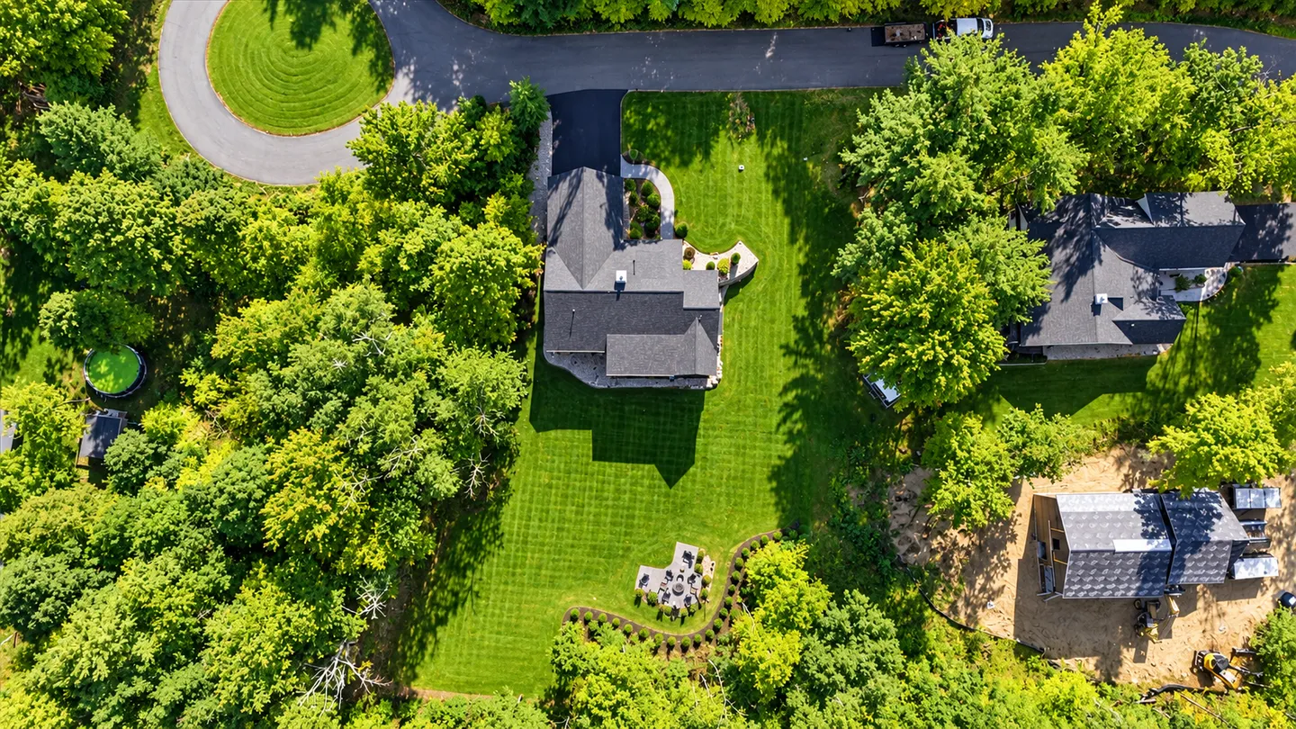 Aerial overhead view of a home surrounded by lush green trees, a large lawn, curved driveway, and a backyard fire pit area.