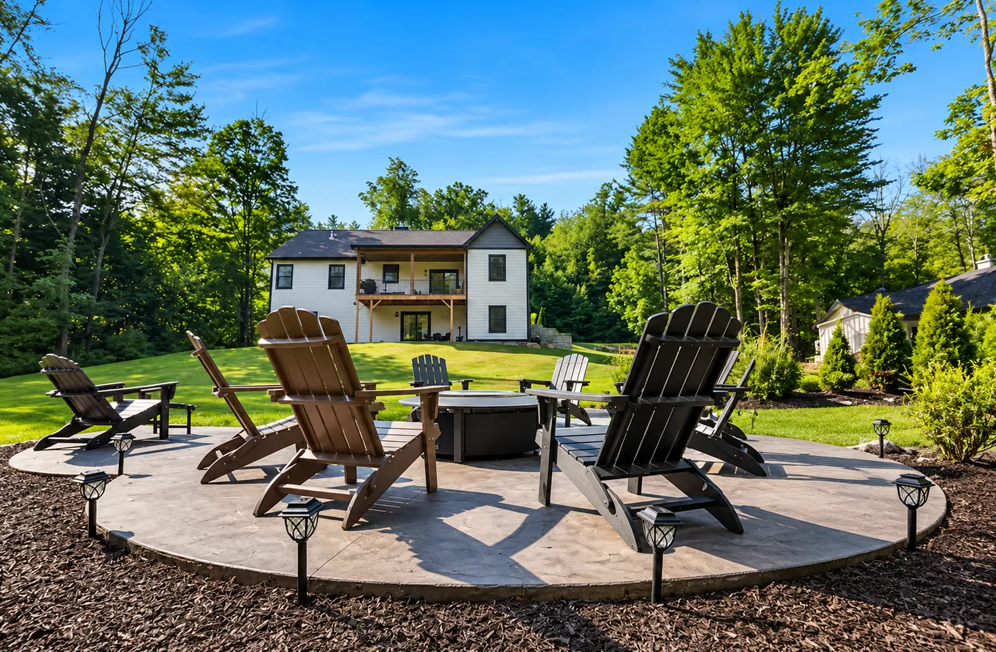 Outdoor circular patio with a fire pit surrounded by Adirondack chairs, overlooking a grassy lawn and two-story house.