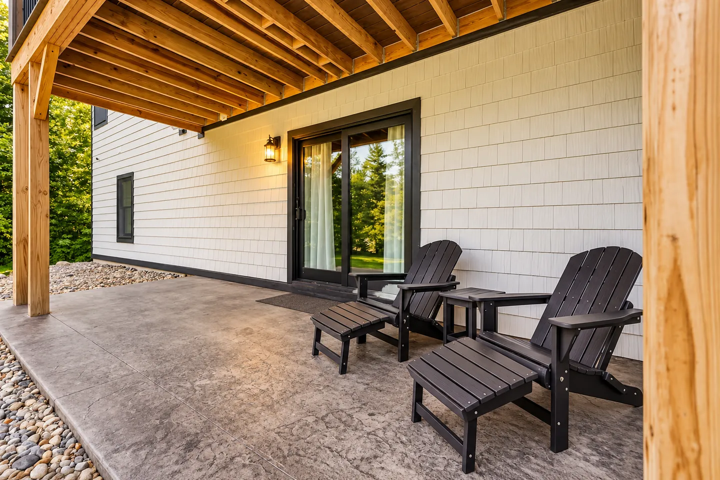 Covered concrete patio under an elevated deck with two dark Adirondack chairs, matching footstools, and a sliding glass door.