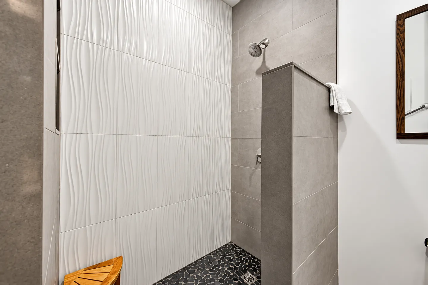 Walk-in shower with textured wave-pattern white wall tiles, gray rectangular tiles, and a pebble-tiled floor.