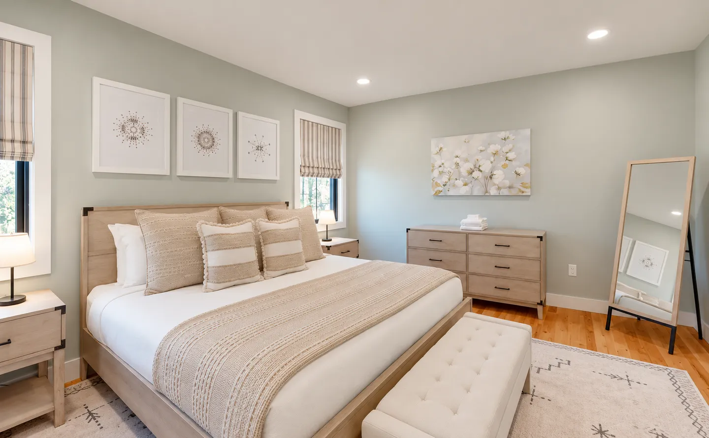 Bedroom with light wooden furniture, a dressed bed with accent pillows, matching dressers, a bench, and a leaning full-length mirror.