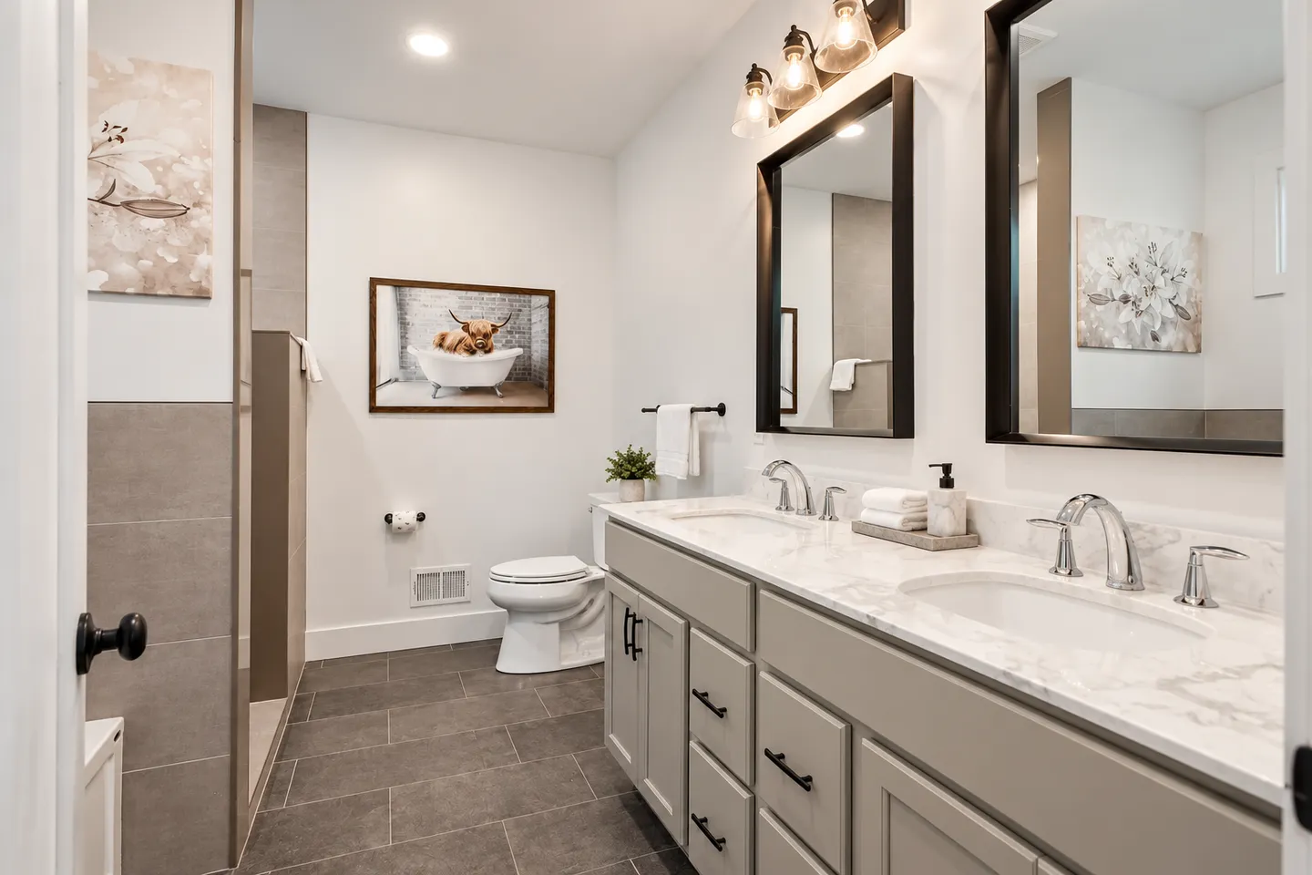 Bathroom with a double-sink vanity, tiled floor, toilet, walk-in shower entrance, and artwork on the walls.