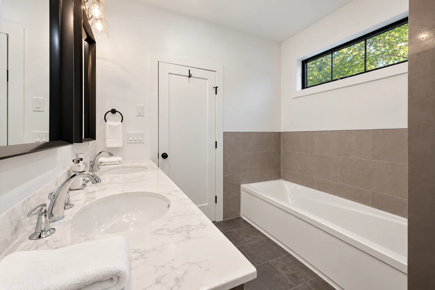 Bathroom featuring a double vanity with marble-patterned countertop and a built-in soaking bathtub beneath a horizontal window.