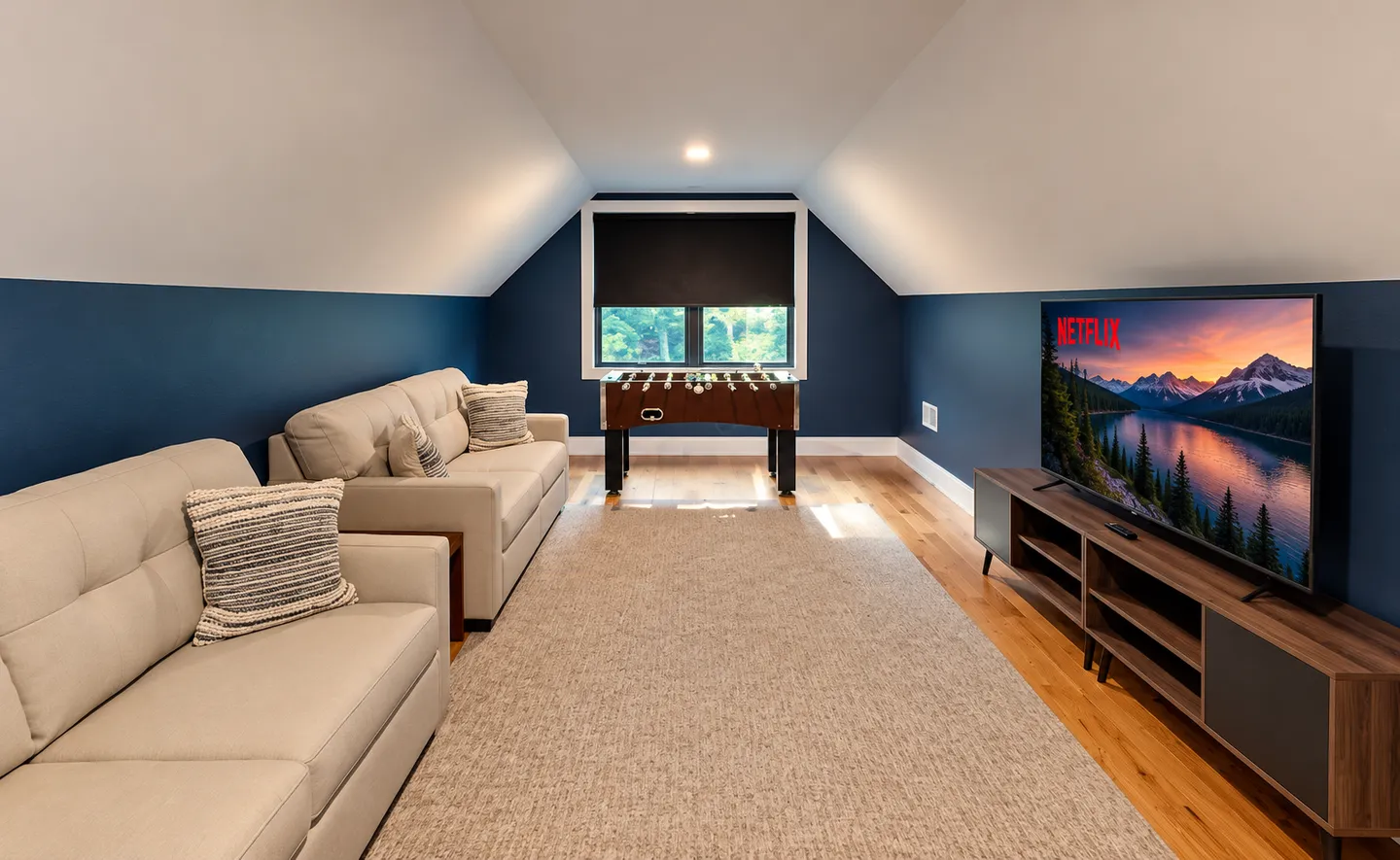Upstairs entertainment room with sloped ceilings, two beige sofas, a foosball table, and a flat-screen TV.