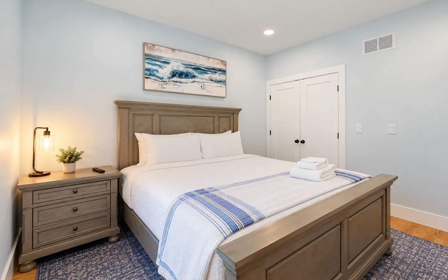 Bedroom with a wooden bed frame, matching nightstand, coastal artwork, and light blue walls.