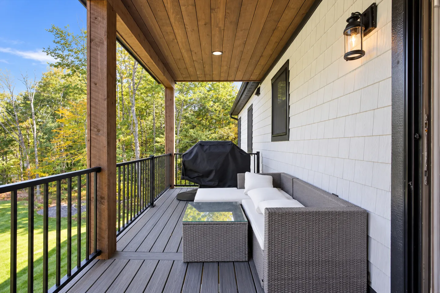 Covered deck with composite flooring, wood-paneled ceiling, outdoor sectional sofa, glass-top coffee table, and a covered barbecue grill.