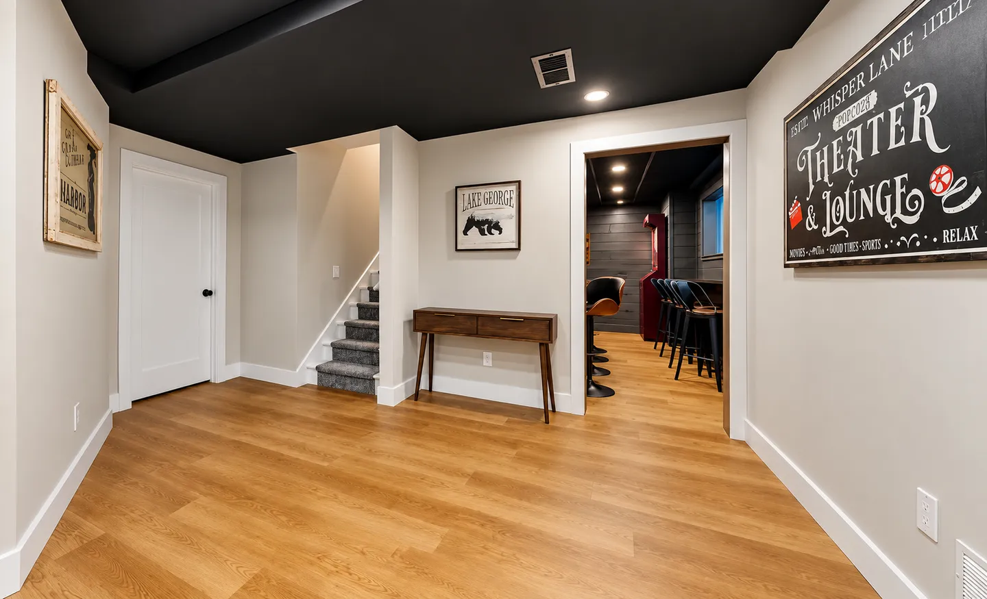 Lower-level foyer with light wood-style flooring, a dark painted ceiling, a staircase with carpet, and a doorway opening to a theater and lounge area.