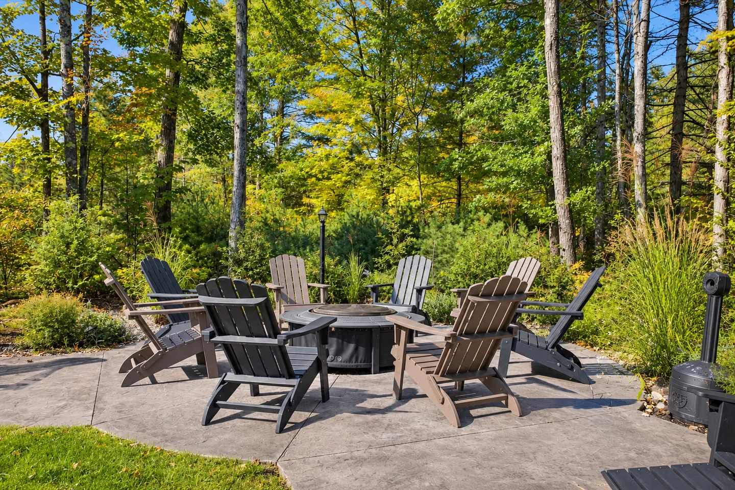 Outdoor concrete patio featuring Adirondack chairs arranged around a central fire pit bordered by lush trees.