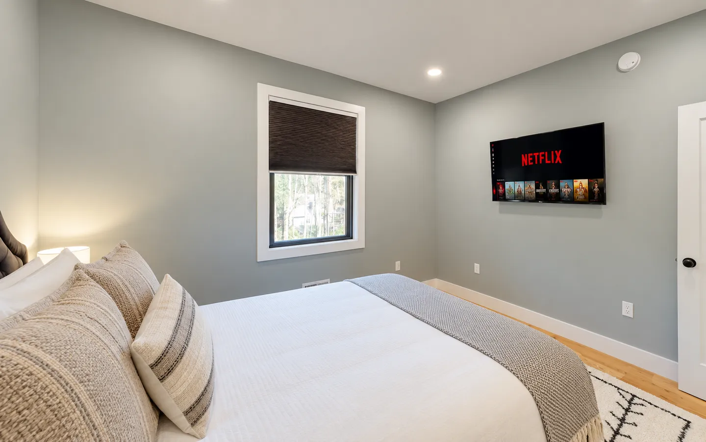 Bedroom featuring a made bed with textured pillows, a wall-mounted flat-screen television, light grey walls, and a window with a dark shade.